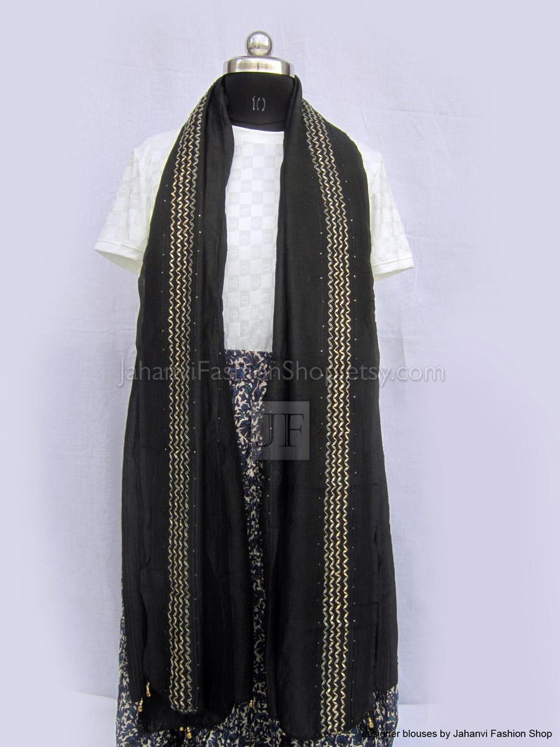 May include: A black scarf featuring two vertical gold zigzag embroidered stripes with small embedded studs. The scarf is draped over a white short-sleeved top with a subtle grid pattern and a blue and white floral print skirt on a mannequin.