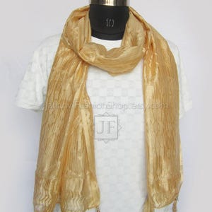 May include: A gold-colored scarf with a textured, pleated design. The scarf is draped around a mannequin and features small tassels at the ends. The scarf is made of a lightweight material.