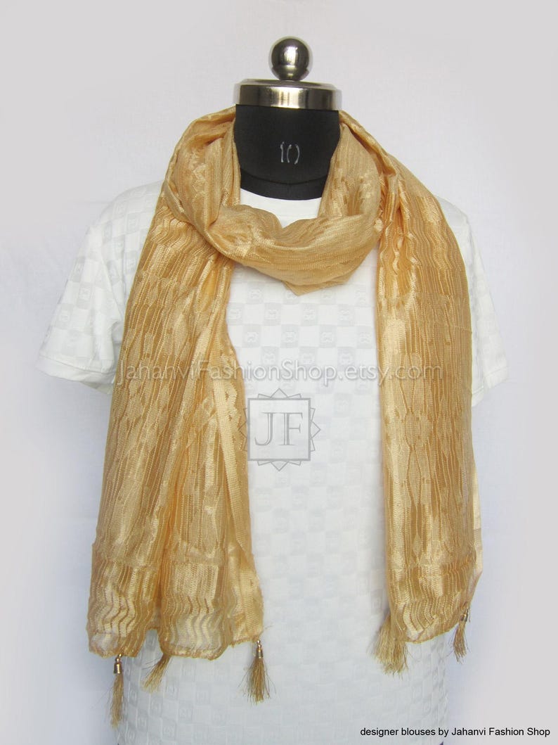 May include: A gold-colored scarf with a textured, pleated design. The scarf is draped around a mannequin and features small tassels at the ends. The scarf is made of a lightweight material.