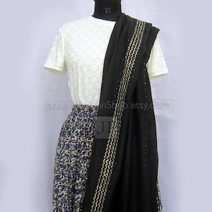 May include: A white short-sleeved top with a subtle checkered texture paired with a long, patterned skirt featuring blue and brown floral motifs. A black shawl with gold zigzag embroidery and small embellishments is draped over the shoulder.