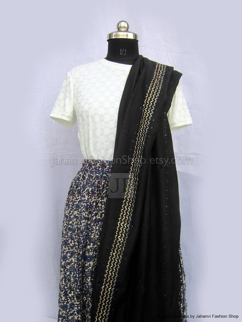 May include: A white short-sleeved top with a subtle checkered texture paired with a long, patterned skirt featuring blue and brown floral motifs. A black shawl with gold zigzag embroidery and small embellishments is draped over the shoulder.