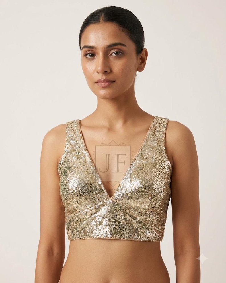 May include: A sleeveless gold sequin crop top featuring a deep V-neckline and a fitted silhouette. The garment is covered in shimmering metallic sequins that reflect light, creating a textured surface. A faint JF logo is visible in the center.