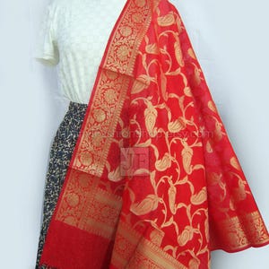 May include: A vibrant red dupatta with gold floral and paisley patterns. The dupatta features intricate gold borders and is draped over a white top and a patterned skirt. The fabric appears lightweight and flowing.