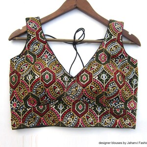 May include: A sleeveless, embroidered top with a deep V-neck. The intricate pattern features red, pink, gold, and green geometric shapes on a black background. The top is hanging on a wooden hanger.