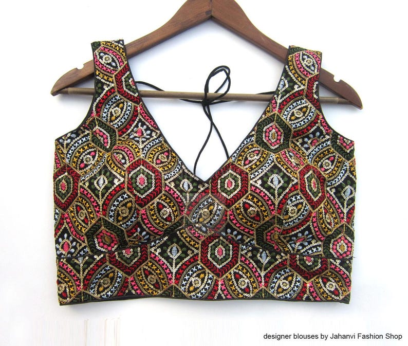 May include: A sleeveless, embroidered top with a deep V-neck. The intricate pattern features red, pink, gold, and green geometric shapes on a black background. The top is hanging on a wooden hanger.