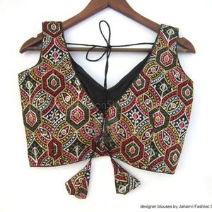 May include: A sleeveless black blouse with a V-neck and intricate embroidery. The blouse features a colorful geometric pattern in red, green, yellow, and white. The blouse is hanging on a wooden hanger.