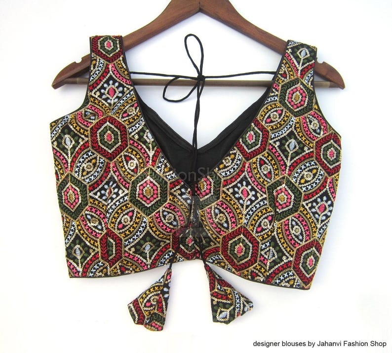 May include: A sleeveless black blouse with a V-neck and intricate embroidery. The blouse features a colorful geometric pattern in red, green, yellow, and white. The blouse is hanging on a wooden hanger.
