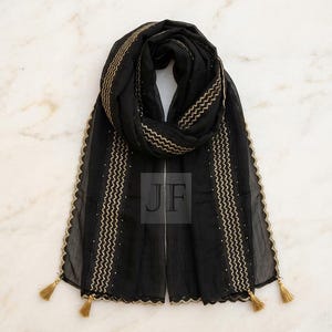 May include: A black scarf featuring gold-colored zigzag embroidery along the vertical edges, a scalloped gold-trimmed border, and small gold tassels at each corner. The center of the scarf displays the letters JF in a square overlay.