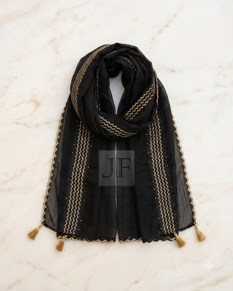 May include: A black scarf featuring gold-colored zigzag embroidery along the vertical edges, a scalloped gold-trimmed border, and small gold tassels at each corner. The center of the scarf displays the letters JF in a square overlay.