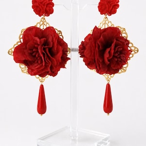 Op de afbeelding: Een paar rode oorbellen met een kleine gevormde rozenknop, een goudkleurig vierkant filigraan frame, een grote stoffen bloem en een gladde rode druppelvormige hanger. De tekst FLAMENCO DANCE SHOP is zichtbaar in de linkerbovenhoek.