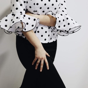 Puede incluir: Un vestido blanco con lunares negros y mangas con volantes. El vestido tiene una falda negra con un dobladillo con volantes.