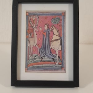 May include: Framed medieval artwork depicting a figure in a blue cloak, gesturing towards a deer and a horse. The artwork has a pink and blue background, and is set in a black frame. The image is a historical illustration.