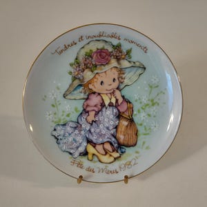 Op de afbeelding: Een rond decoratief porseleinen bord met een gouden rand, voorzien van een illustratie van een kind met een bloemenhoed, een paarse jurk en gele schoenen. De tekst luidt Tendres et inoubliables moments en Fête des Mères 1982.