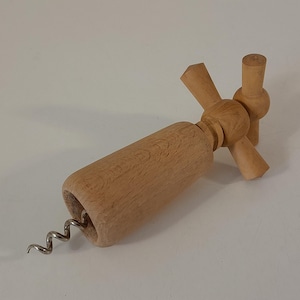 Könnte beinhalten: Ein hellfarbener Holzkorkenzieher mit einem zylindrischen Griff und einem T-förmigen Drehmechanismus an der Oberseite. Eine silberfarbene Metallspirale ragt aus der Basis des Holzkörpers heraus und dient zum Herausziehen von Weinkorken.