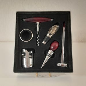 Op de afbeelding: Een wijnaccessoireset gepresenteerd in een zwarte doos met schuimvoering, bestaande uit een T-vormige kurkentrekker met een donkerrood handvat, een zilveren druppelring, een metalen schenker, een flesstop met een donkerrode bovenkant en een thermometer.