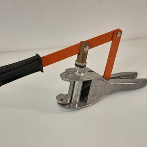 Op de afbeelding: Een handmatig metalen zeilringpersgereedschap met een zilverkleurige basis waarop het woord "SAMPEI" in reliëf staat, een oranje hefboomarm en een zwarte kunststof handgreep met het woord "SAMPEI" in het oppervlak van de greep gegoten.