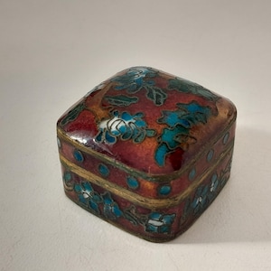Pode incluir: Uma pequena caixa quadrada de metal com trabalho de esmalte cloisonné sobre um fundo vermelho profundo com padrões florais vibrantes em azul e verde. A tampa e os lados são decorados com contornos de fio metálico e as bordas têm um acabamento em latão.