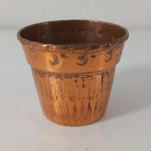 Op de afbeelding: Een kleine, metalen koperkleurige plantenpot met een taps toelopende cilindrische vorm. De bovenrand is voorzien van een decoratieve band met reliëf in de vorm van maansikkels, terwijl het onderste deel een textuur met een verticaal geborstelde afwerking heeft.