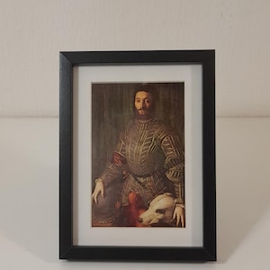 Puede incluir: Impresión enmarcada de un retrato renacentista. El sujeto, un hombre barbudo, viste un jubón estampado y sostiene un perro. El marco negro rodea la impresión, que está sobre un passe-partout blanco.