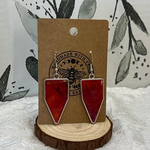 May include: A pair of handmade earrings featuring red and orange resin in a teardrop shape. The earrings are silver and have a small bee charm on the hook.
