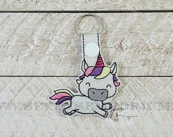May include: A white unicorn keychain with a pink, purple and yellow mane and tail. The unicorn is wearing a purple and pink party hat. The keychain is on a wooden background.
