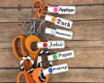 Puede incluir: Un juego de seis tijeras con mango naranja y etiquetas de diferentes colores. Las etiquetas están etiquetadas como "Aplique", "Pato", "Hemostáticos", "Tela", "Papel" y "Punteado".