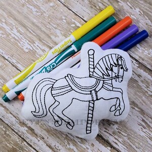 May include: A white fabric carousel horse with black outlines, ready to be coloured in. The horse is lying on a wooden surface with a set of colourful markers beside it.