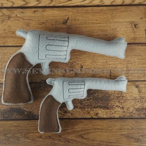 May include: Two grey felt toy guns with brown felt handles. The guns are lying on a wooden surface.