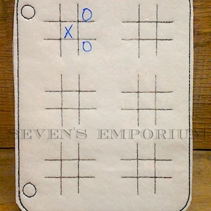 以下が含まれることがあります： 黒いステッチと青いマークが付いた白い布の三目並べボード。ボードには三目並べのグリッドが3つあります。上のグリッドには「X」と「O」が2つあります。ボードの下部には「SEVEN'S EMPORIUM」というテキストが表示されています。