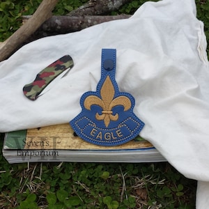May include: A blue and gold embroidered patch with the word "EAGLE" and a fleur-de-lis design. A camouflage pocket knife rests on a white cloth. The patch is on top of a book with the text "Seven's Emporium".