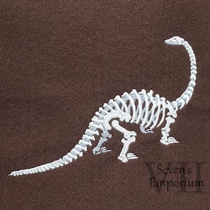 以下が含まれることがあります： 茶色の布地に白い刺繍で描かれた恐竜の骨格。恐竜は四足で立ち、頭を上げています。右下には「Seven's Emporium」の文字が見えます。