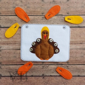 Può includere: Un'attività di artigianato in feltro con un tacchino di feltro marrone con testa gialla e bargiglio rosso. Il tacchino ha bottoni di feltro marrone per le piume. Ci sono sei pezzi di feltro arancio e giallo da posizionare sul tacchino per completare l'artigianato.