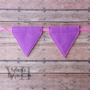 Könnte beinhalten: Zwei violette Filz-Dreiecksfahnen mit weißen Nähten, die an einem rosa Band mit weißen Punkten hängen. Die Fahnen sind leer und können personalisiert werden.