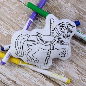 May include: A white fabric carousel horse with black embroidery, ready to be coloured in with markers. The horse is on a wooden surface with markers in various colours.