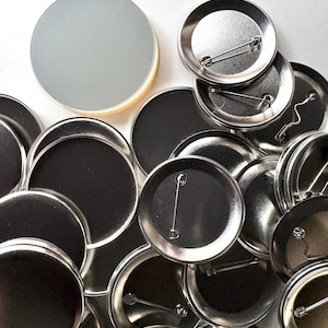 May include: A collection of blank, round metal pinback buttons. The buttons have a shiny silver finish on the front and a black back. Some buttons are face up, showing the pin and clasp.