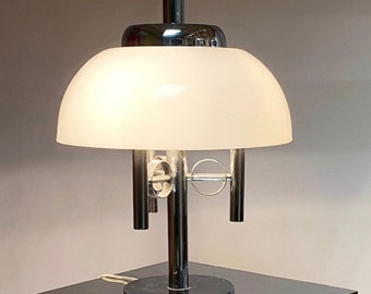 May include: A chrome table lamp with a white frosted glass shade and a unique design featuring multiple chrome rings. The lamp has a round base and a tall, slender stem.