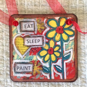 May include: Square, mixed-media art piece with a brown border and red ribbon. The artwork features the words "EAT," "SLEEP," and "PAINT" in rectangular blocks. Yellow and blue flowers are painted on the right side, with a red and white background.