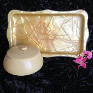 May include: A vintage serving set featuring a rectangular tray and a lidded container. The tray and container are a creamy yellow color with a geometric design. The set is displayed on a black velvet surface.