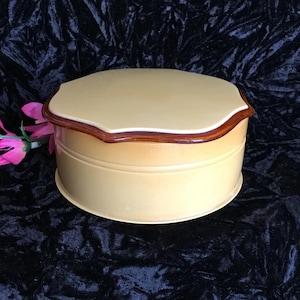 May include: A round, vintage jewelry box with a light yellow body and a scalloped lid. The lid is trimmed with a dark brown border. The box sits on a black velvet surface. A pink flower is visible in the background.
