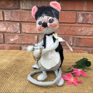 May include: A gray plush mouse wearing a tuxedo and holding a silver trophy. The mouse has black hair, pink ears, and a white flower on its lapel. The mouse is standing on a brown burlap surface.