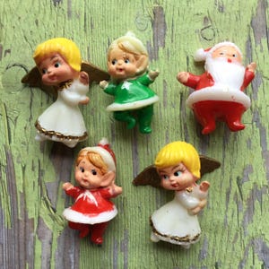 May include: A collection of vintage Christmas figurines, including angels with gold wings, a Santa Claus, and an elf. The figurines are in various colors, including red, white, green, and yellow, and are arranged on a weathered green surface.