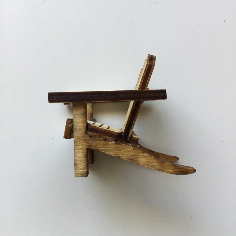 May include: A miniature wooden adirondack chair with a brown stained seat and back. The chair is made of light wood and has a simple design.