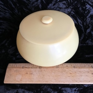 May include: A vintage, cream-colored, lidded jar with a rounded body and a small knob on the lid. The jar is made of a smooth, solid material. A wooden ruler with inch markings is placed below the jar.
