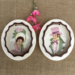 May include: Two vintage ceramic wall plates with scalloped edges and a dark brown trim. One plate features a woman in a hat, the other a man in a top hat. Both are painted in pastel colors, with a floral accent.