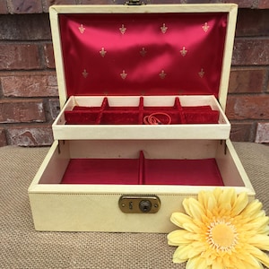 May include: Open vintage jewelry box with a cream-colored exterior and a red velvet interior. The box has a divided tray and a lower compartment. A yellow flower is in the foreground.