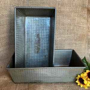 May include: Two rectangular metal loaf pans with a textured dimpled surface. The upright pan features the embossed text OVENEX THE NEW BAKING METAL PAT NO 2077757. These silver-toned baking pans have a worn, metallic finish and straight sides.