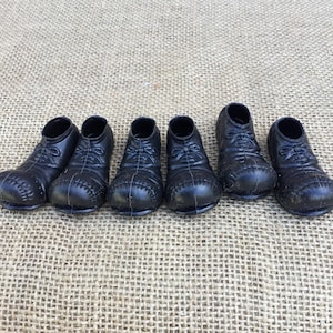 May include: Six small, black toy shoes are arranged in a row. The shoes have laces and a rounded toe. The shoes are made of a shiny material and are set against a textured, tan background.