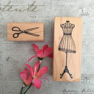 May include: Two wooden stamps with black ink designs. One features scissors, the other a dressmaker's form. Pink flowers are in the foreground. The background is a collage of vintage newspaper clippings.