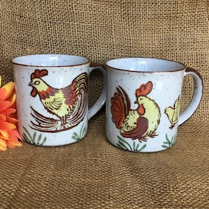May include: Two ceramic mugs with a speckled white finish and brown handles. Each mug features a rooster illustration in yellow, brown, and red, with green grass accents. The mugs are suitable for coffee or tea.