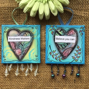 May include: Two square, hand-painted art pieces with a heart design. One says "Kindness Matters" and the other says "Believe you can". Each has a blue border, a ribbon for hanging, and dangling crystal beads. The background is a burlap texture.